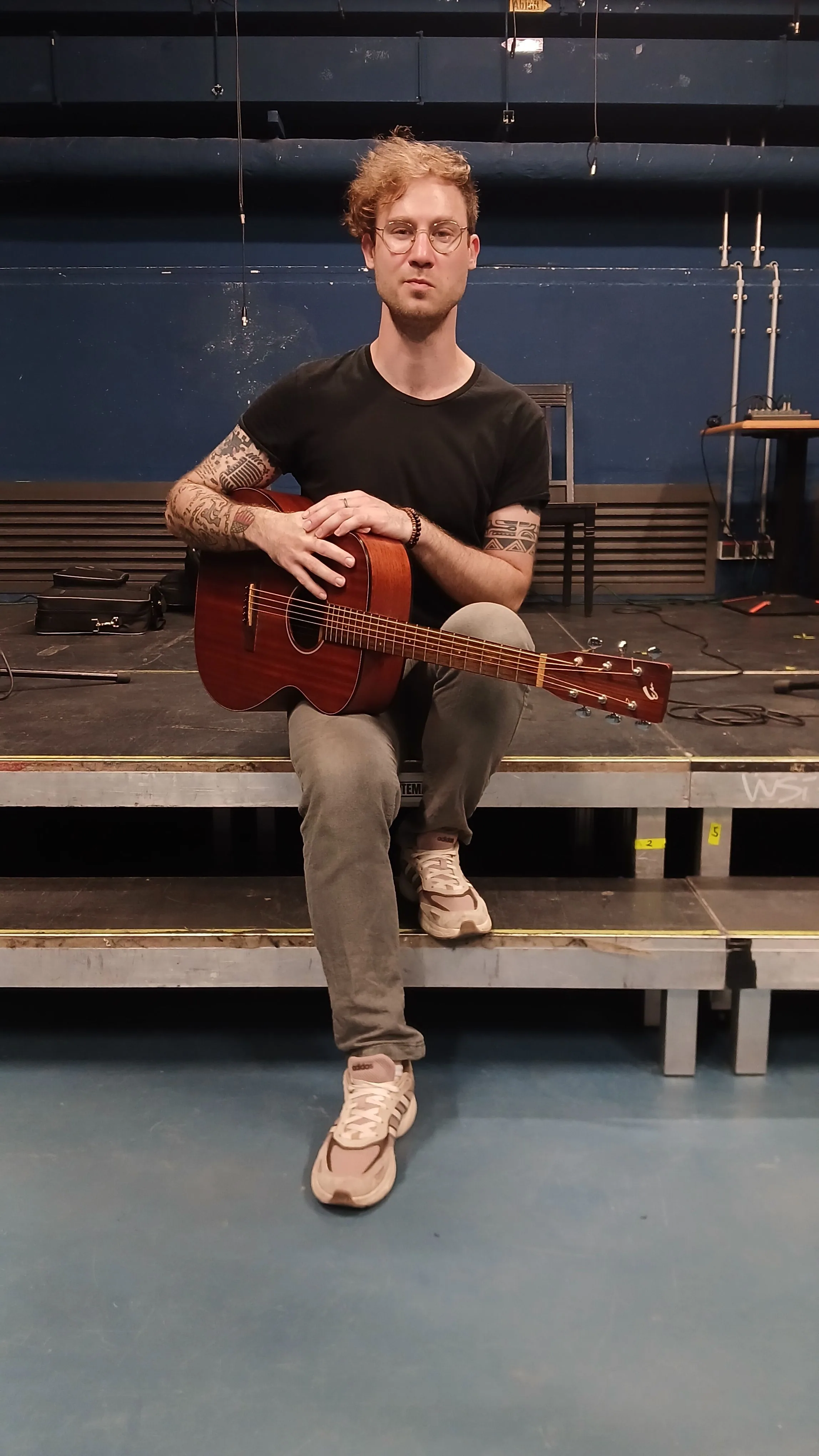 Bricky Waters hält seine Gitarre und blickt in die Kamera, Bühne im Hintergrund